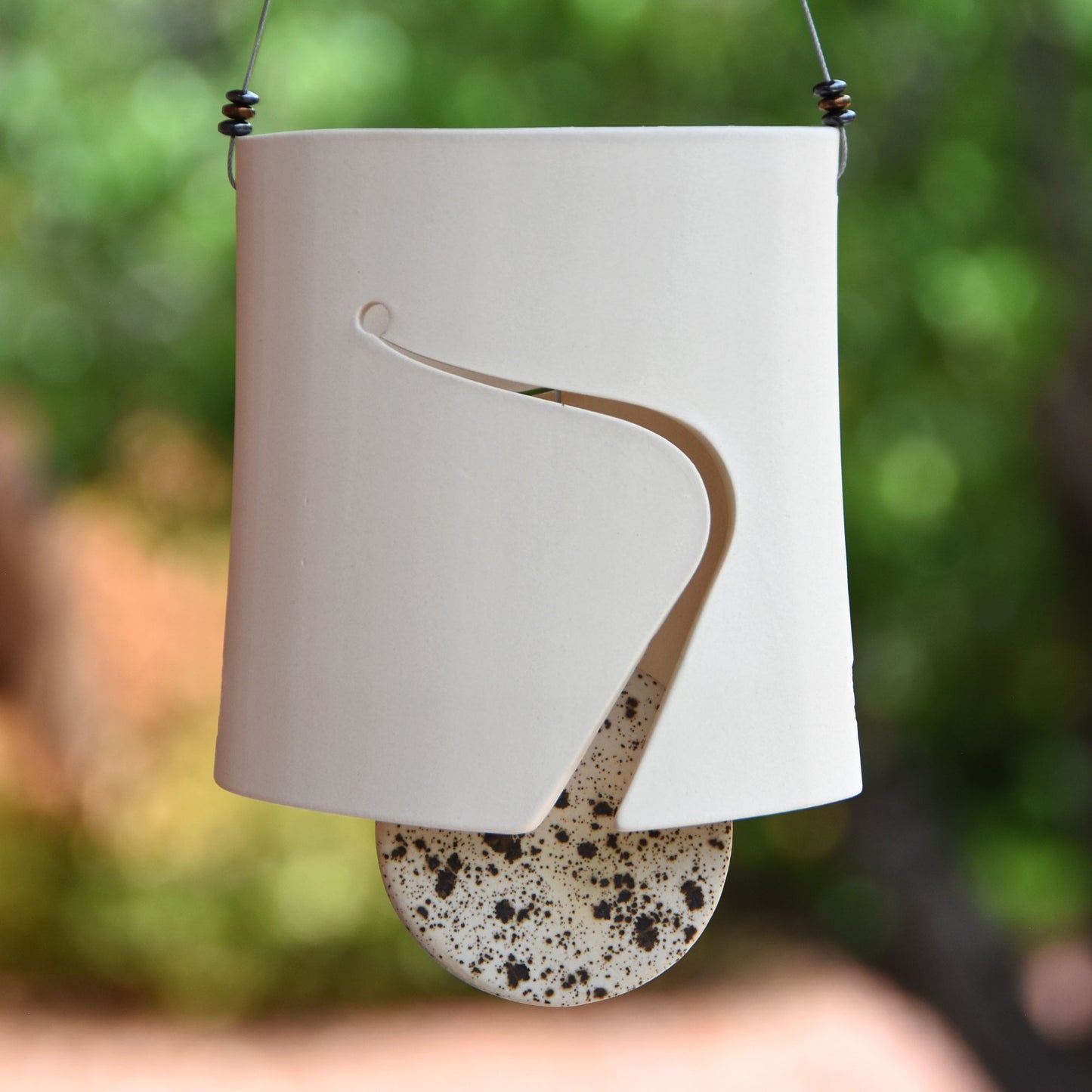 Minimalist white ceramic wind chime hanging from steel wire which is adorned with three modern steel beads on each side and wind chime body has an artistic cut for sound hole and a speckled black and white ceramic clapper