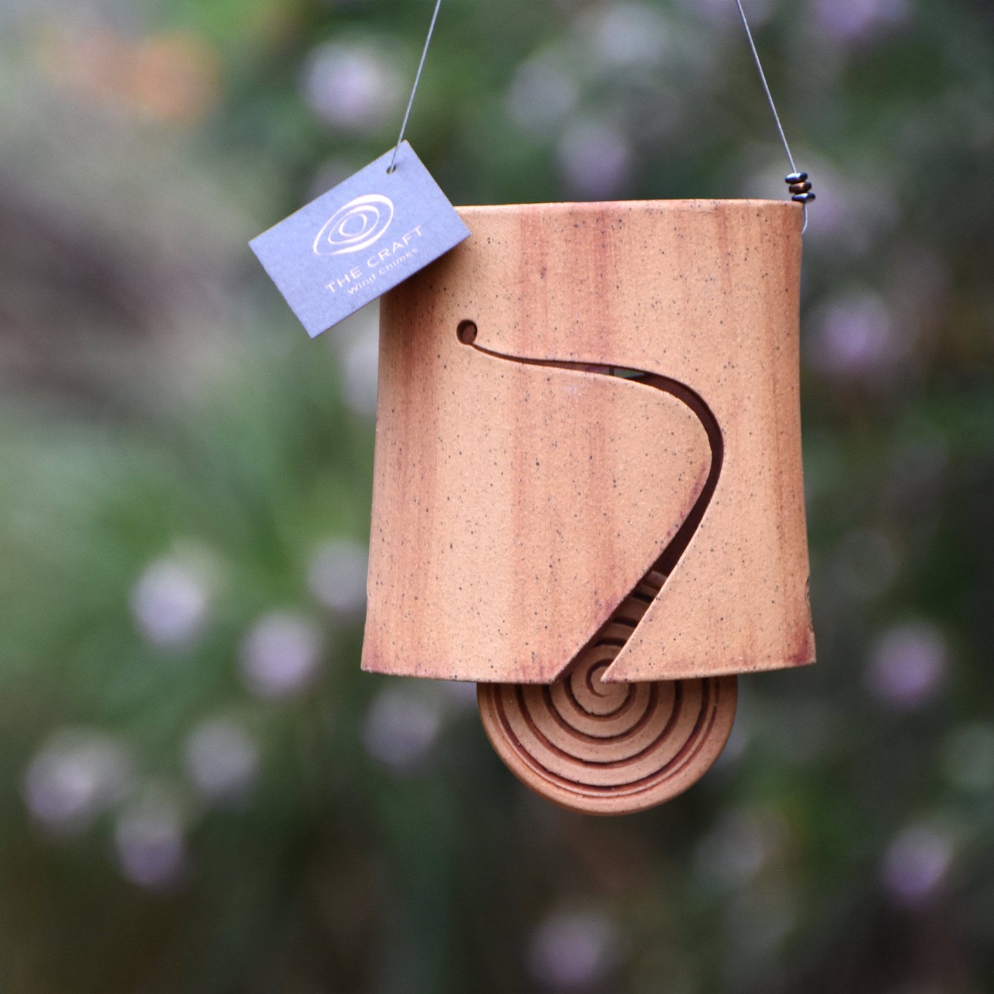 Modern speckled buff ceramic wind chime bell that is stained with red iron oxide. The bell features a round spiral carved wind clapper, also stained with red iron oxide. The bell hangs from a stainless steel wire which is adorned with steel beads.