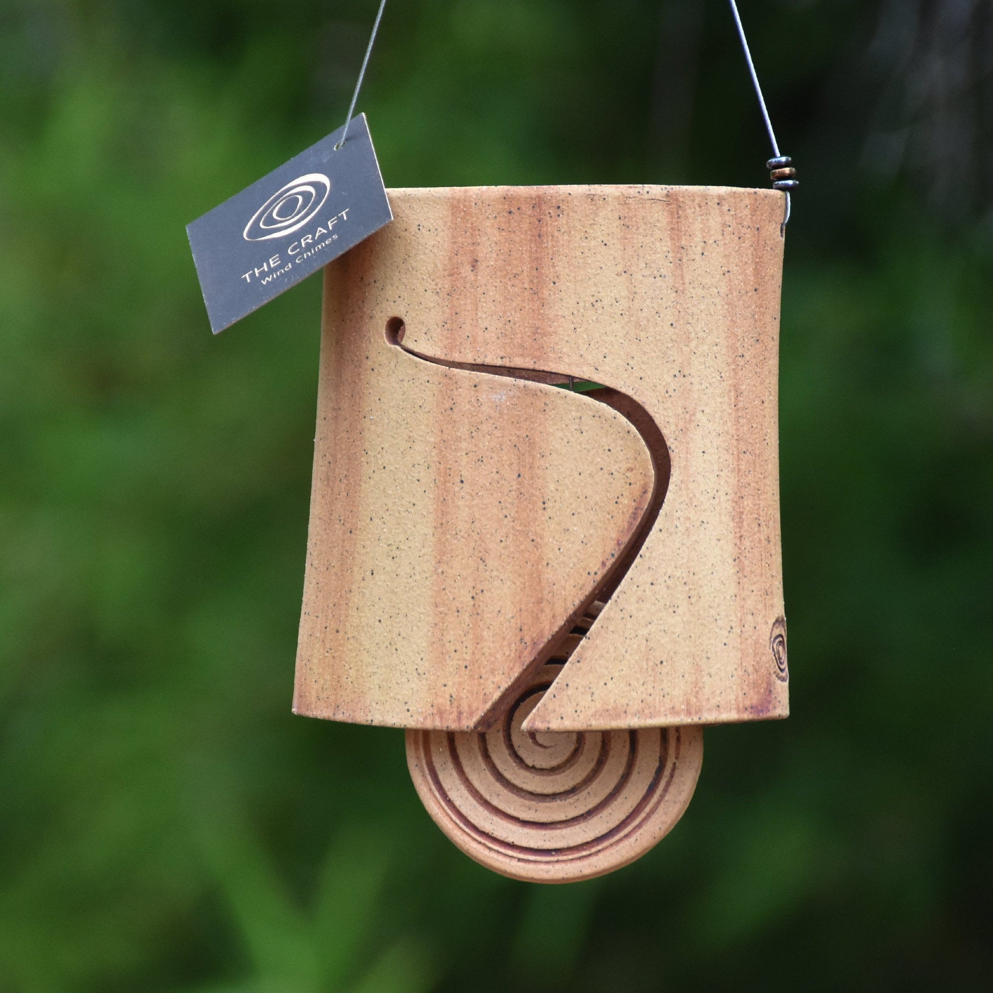 Modern speckled buff ceramic wind chime bell that is stained with red iron oxide. The bell features a round spiral carved wind clapper, also stained with red iron oxide. The bell hangs from a stainless steel wire which is adorned with steel beads.