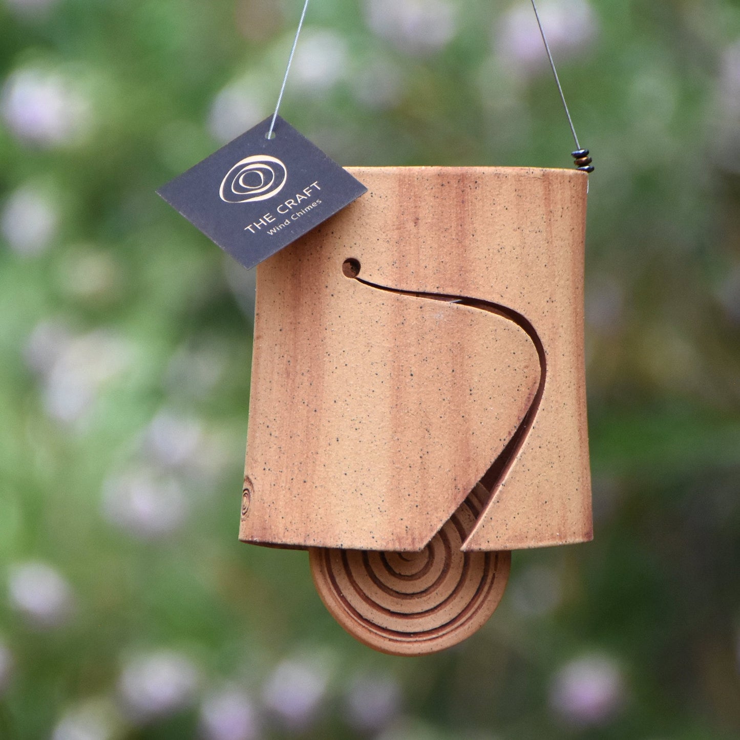 Modern speckled buff ceramic wind chime bell that is stained with red iron oxide. The bell features a round spiral carved wind clapper, also stained with red iron oxide. The bell hangs from a stainless steel wire which is adorned with steel beads.