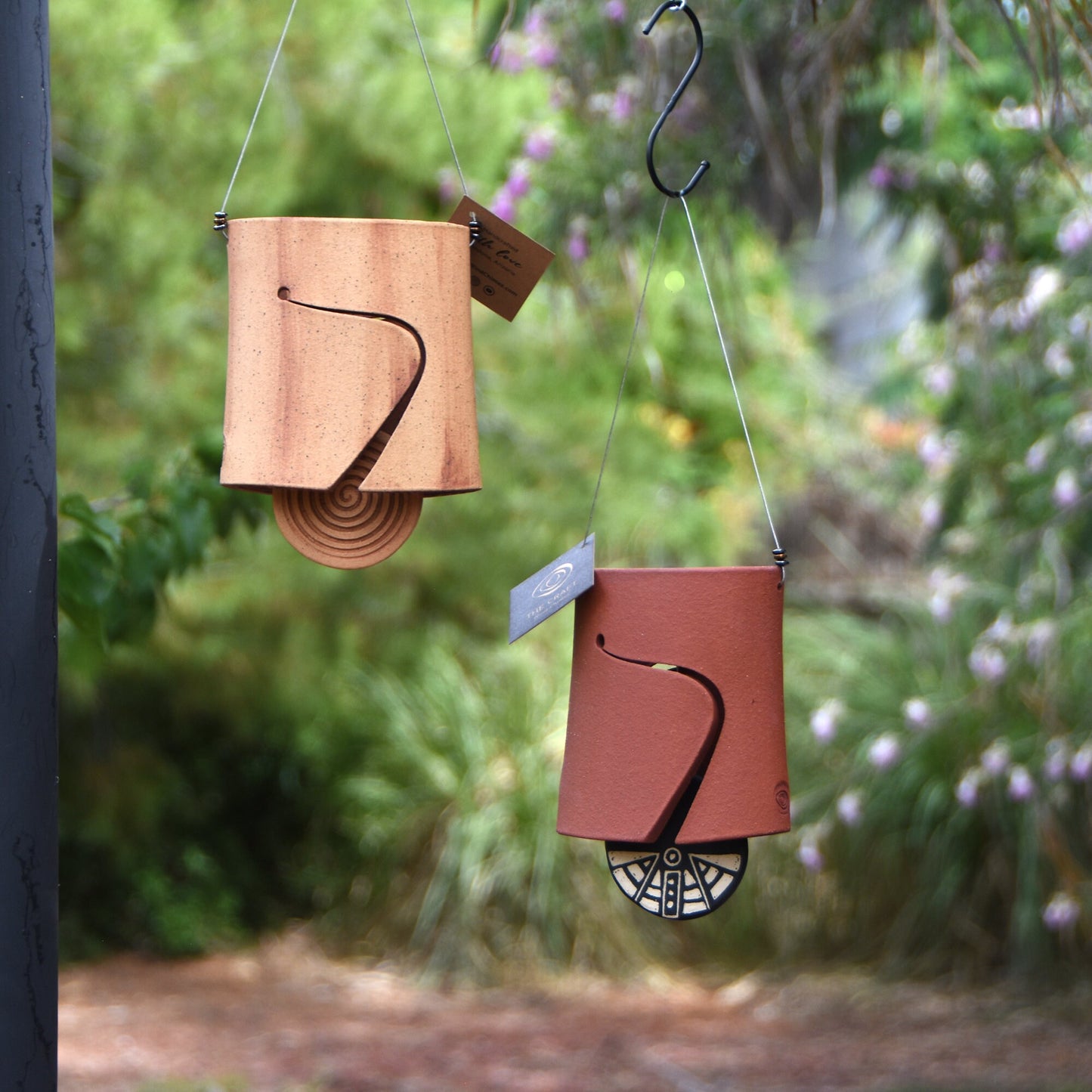 Modern speckled buff and modern rustic red ceramic wind chime bells that were stained with red iron oxide. One bell shows a spiral clapper, the other shows a black and white sgraffito wind clapper. Both are hanging from a stainless steel wire