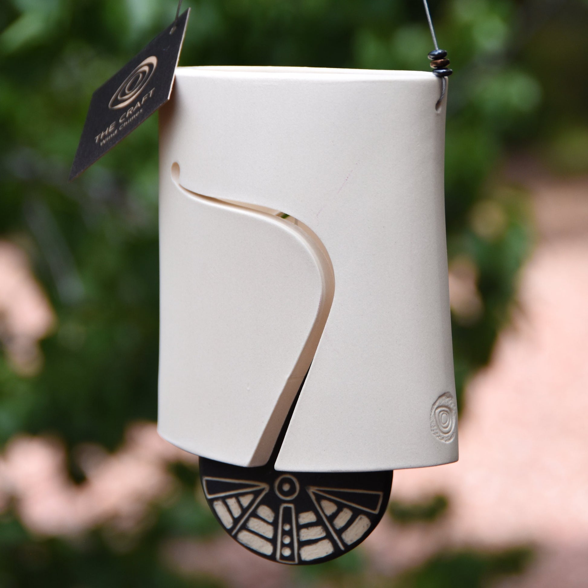 Modern white wind chime bell with sound holes carved on both sides of the oval-shaped body. Bell features a black and white carved wind clapper with an abstract motif on both sides. Bell hangs from a stainless steel wire, adorned with steel beads.