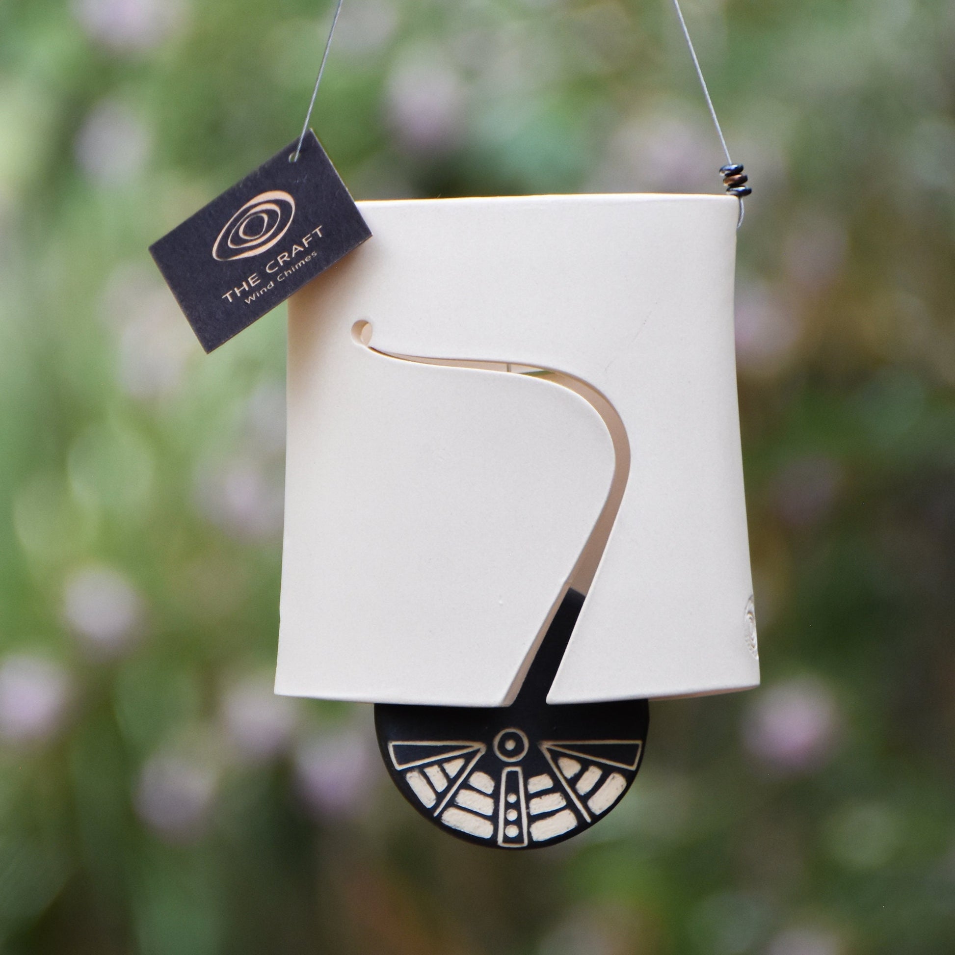 Modern white wind chime bell with sound holes carved on both sides of the oval-shaped body. Bell features a black and white carved wind clapper with an abstract motif on both sides. Bell hangs from a stainless steel wire, adorned with steel beads.