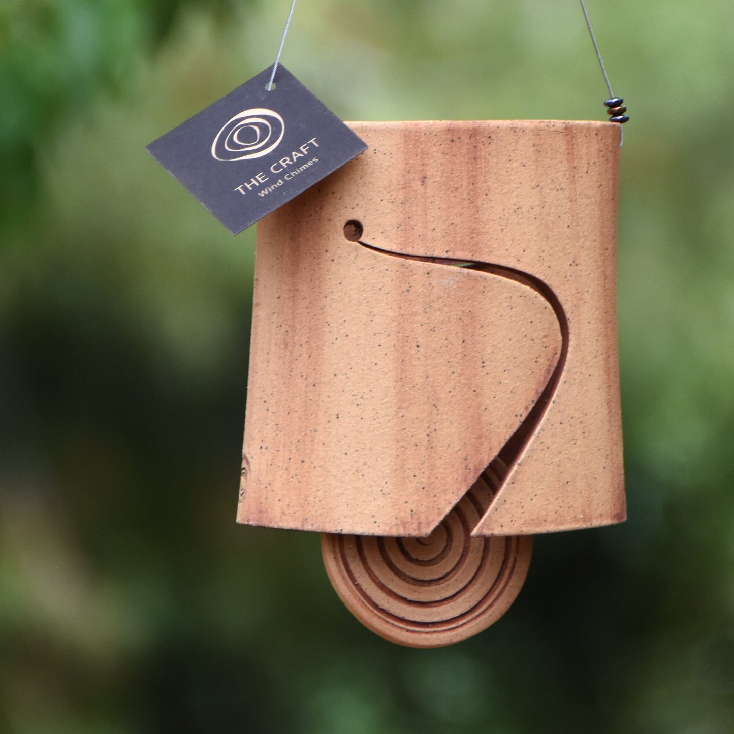 Modern speckled buff ceramic wind chime bell that is stained with red iron oxide. The bell features a round spiral carved wind clapper, also stained with red iron oxide. The bell hangs from a stainless steel wire which is adorned with steel beads.