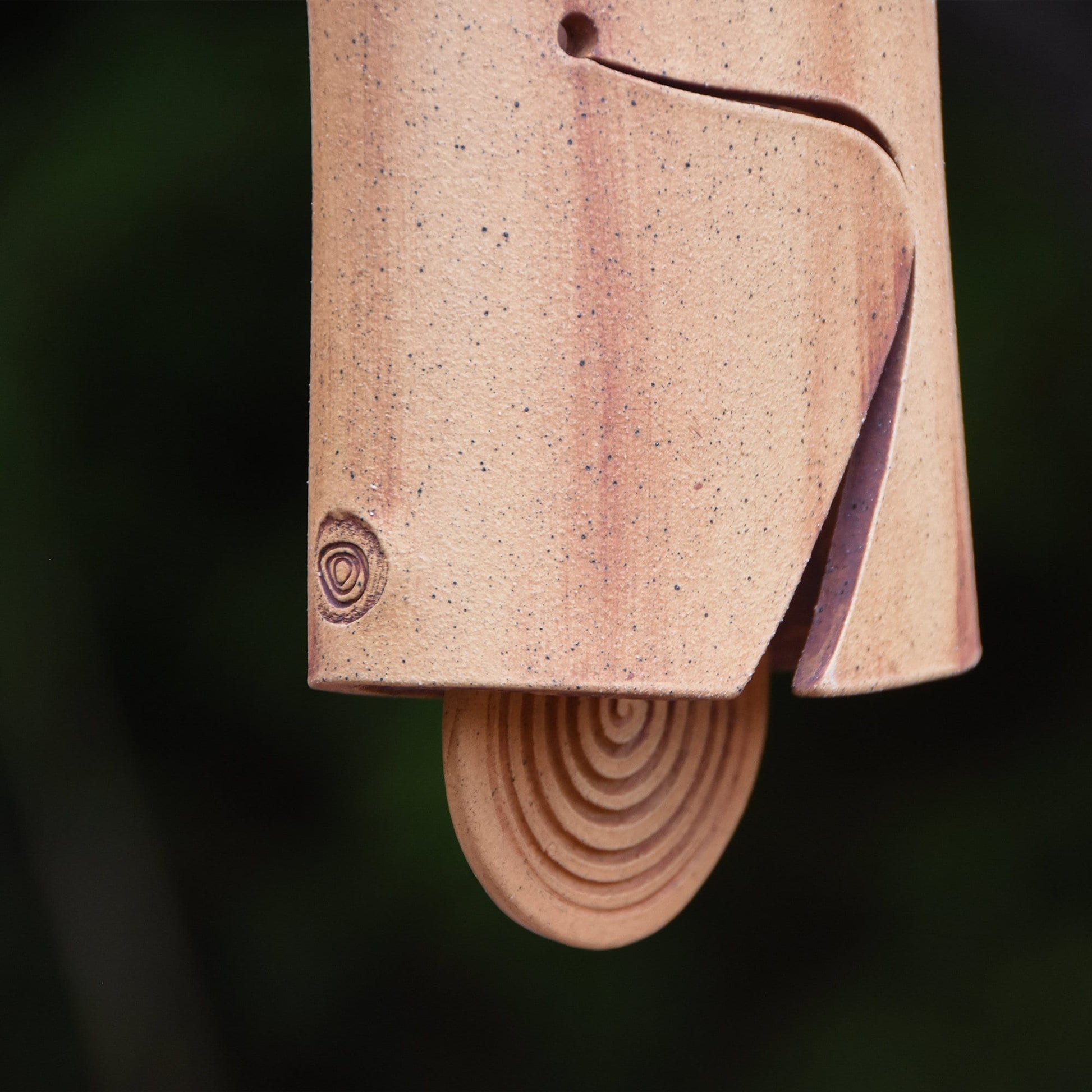 Side view of modern speckled buff ceramic wind chime bell that is stained with red iron oxide. The bell features a round spiral carved wind clapper, also stained with red iron oxide. The side view shows the Craft logo stamped into the clay body