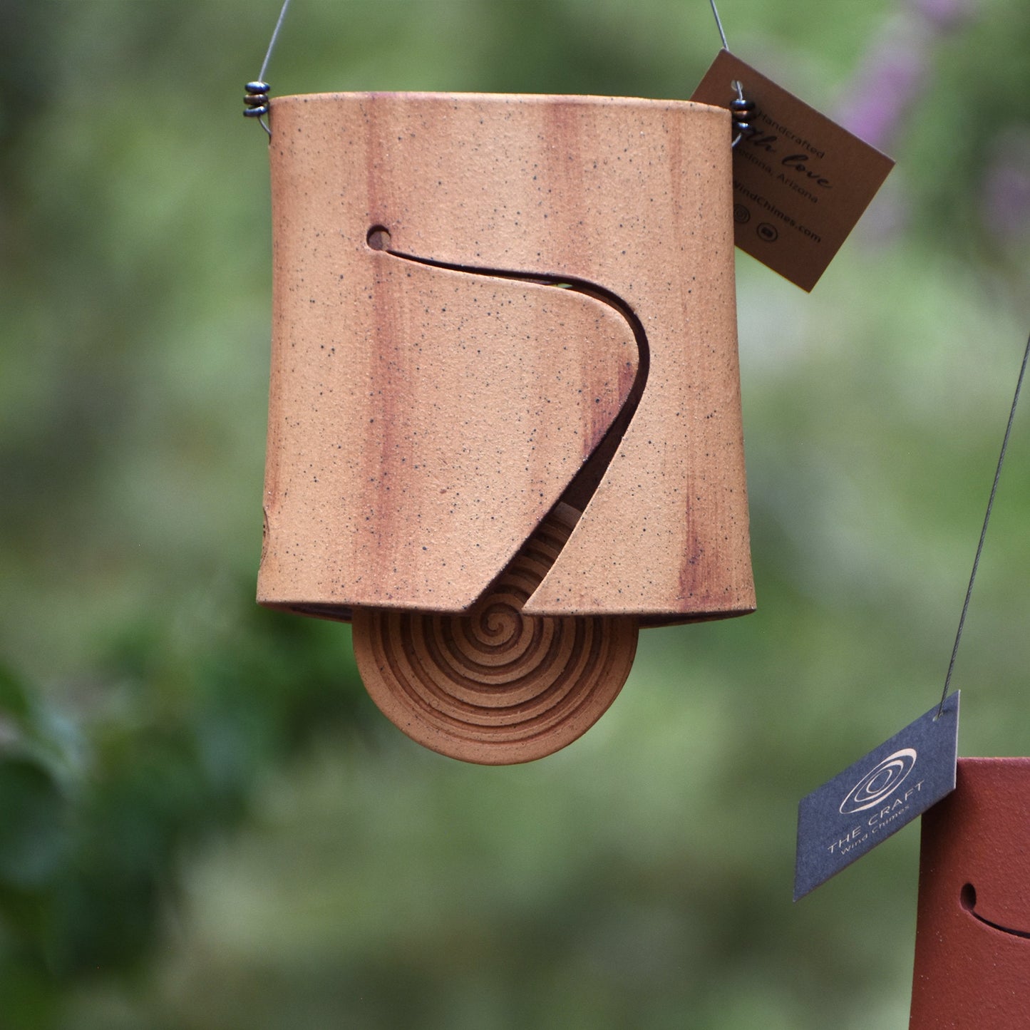 Modern speckled buff ceramic wind chime bell that is stained with red iron oxide. The bell features a round spiral carved wind clapper, also stained with red iron oxide. The bell hangs from a stainless steel wire which is adorned with steel beads.