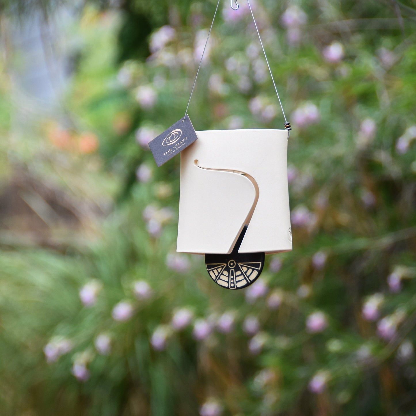 Modern white wind chime bell with sound holes carved on both sides of the oval-shaped body. Bell features a black and white carved wind clapper with an abstract motif on both sides. Bell hangs from a stainless steel wire, adorned with steel beads.